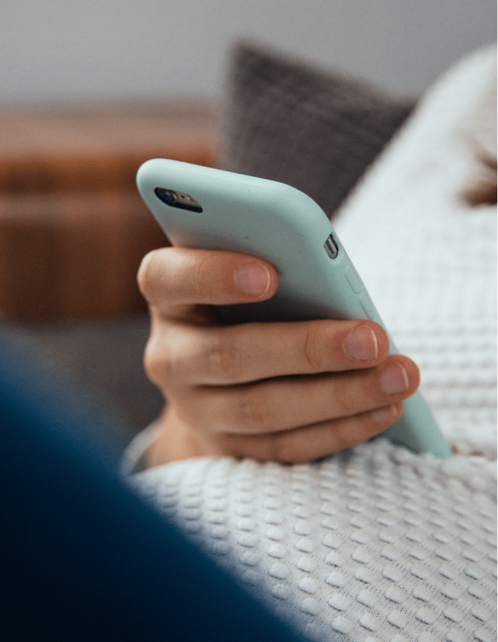 Close-up of a person's hand holding a smartphone with a light blue case while sitting on a textured white blanket.