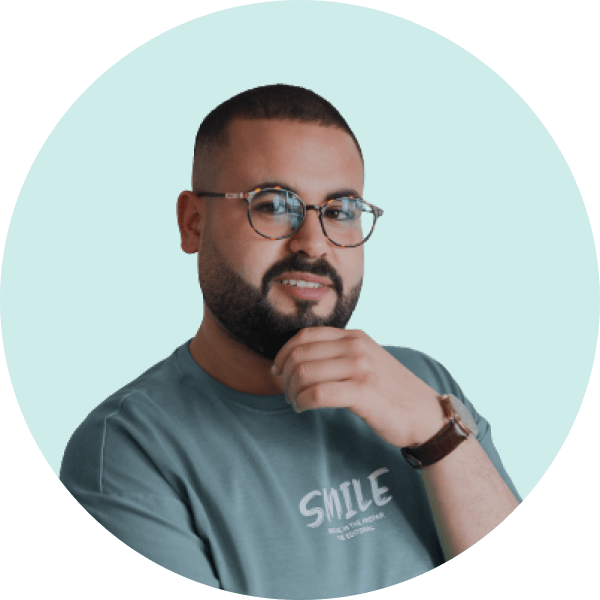 Man with short hair, beard, and glasses resting his chin on his hand, wearing a teal shirt with the word 'SMILE'.