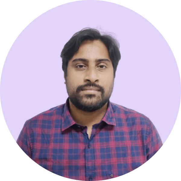 Portrait of a man with dark hair and beard wearing a red and blue checkered shirt against a light purple background.