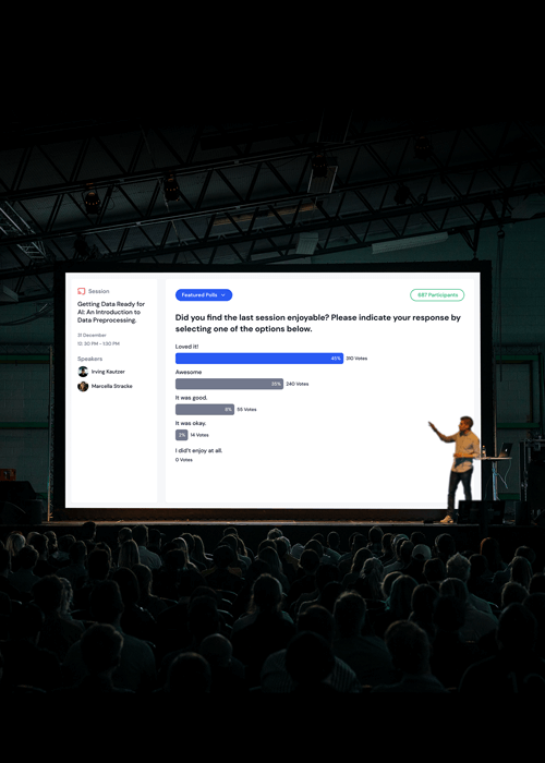 A presenter beside a large screen showing a session feedback poll about enjoyment with options and vote percentages, in a dark auditorium filled with seated audience.