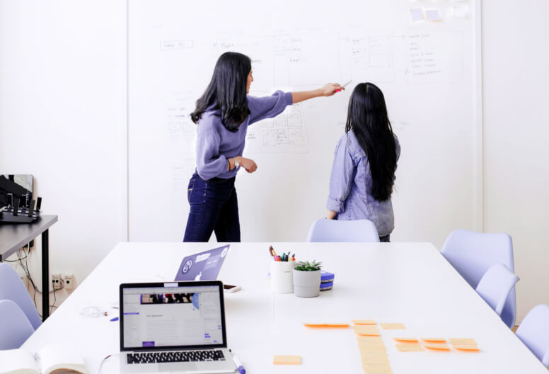 Two women discussing and pointing at notes on a whiteboard in a bright office meeting room with a table, laptop, and stationery.