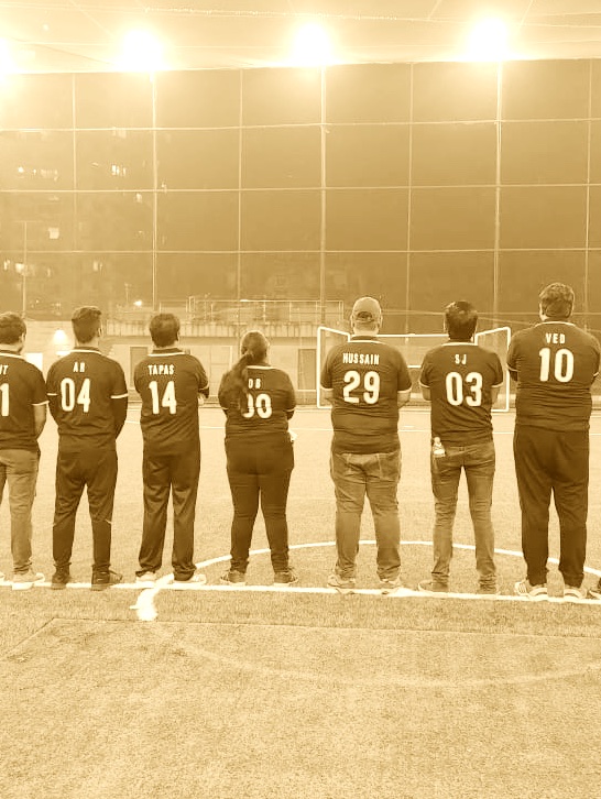 Seven people standing side by side on a sports field at night, wearing matching shirts with names and numbers on the back facing away from the camera.