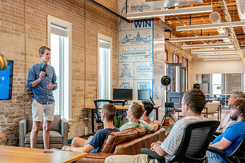Man giving a presentation to a group of people seated in a modern office with exposed brick walls and large windows.