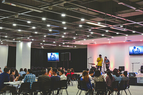 People seated at tables in a large room watching two speakers on stage with microphones and a drum set.
