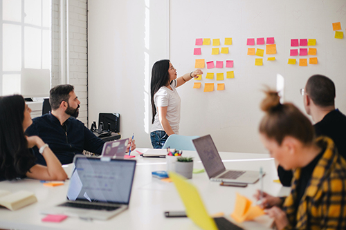 Woman placing sticky notes on a whiteboard during a group meeting with colleagues around a table using laptops and notebooks.