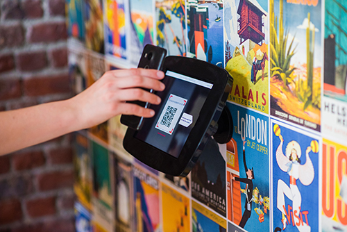 Hand holding a smartphone scanning a QR code on a digital reader mounted on a colorful travel poster wall.