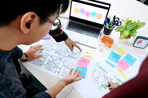 Two people collaborating on user interface design sketches with sticky notes and a laptop on a desk.