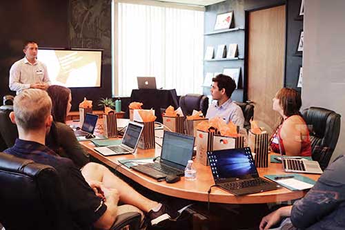 A group of people attending a business presentation in a conference room with laptops and gift bags on the table.
