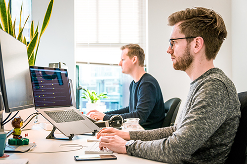 Two men working on laptops at a white desk in a bright office.