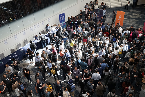 Large group of people mingling in a conference or event lobby with tables and informational signs.