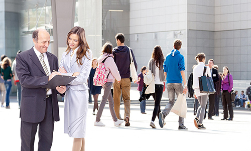 Two business professionals reviewing documents while people walk by outside a modern office building.