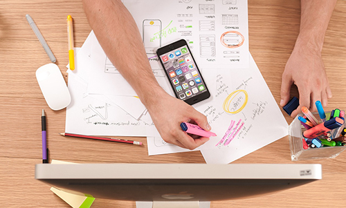 Person annotating printed charts and notes with a pink highlighter at a desk with a smartphone, pens, and a computer monitor.