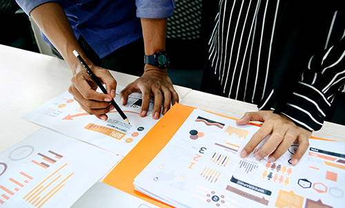 Two people reviewing and marking printed charts and graphs on a white table during a meeting.