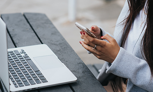 Person with red nail polish using a smartphone beside a laptop on a dark wooden bench.
