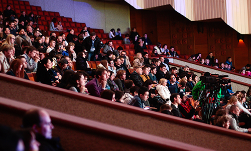 Audience seated in a theater auditorium watching an event with cameras set up on the right side.