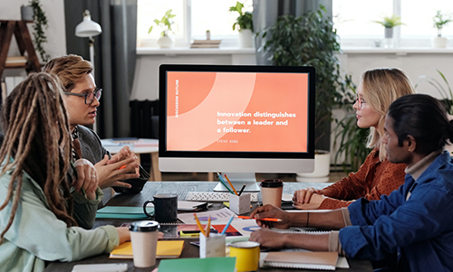 Four people having a meeting around a table with a computer screen showing a quote about innovation and leadership.