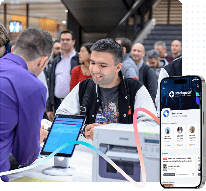 Smiling man talking to a staff member at a registration desk with a digital screen and a smartphone displaying the Startupcon conference app.