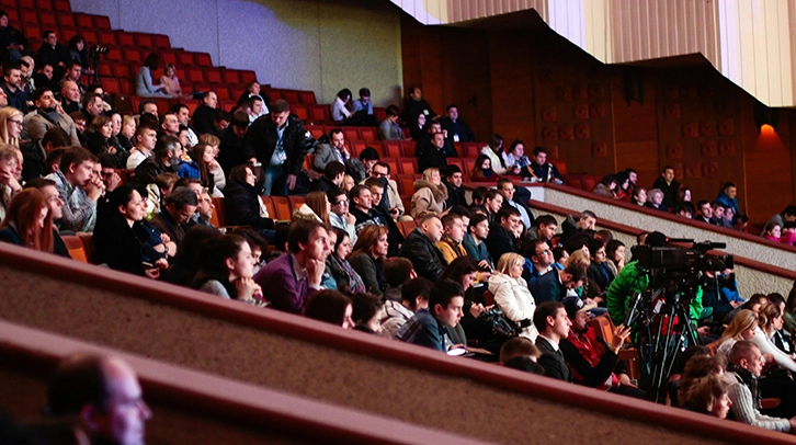 Audience seated in a large auditorium, attentively watching an event, with a few camera operators among them.