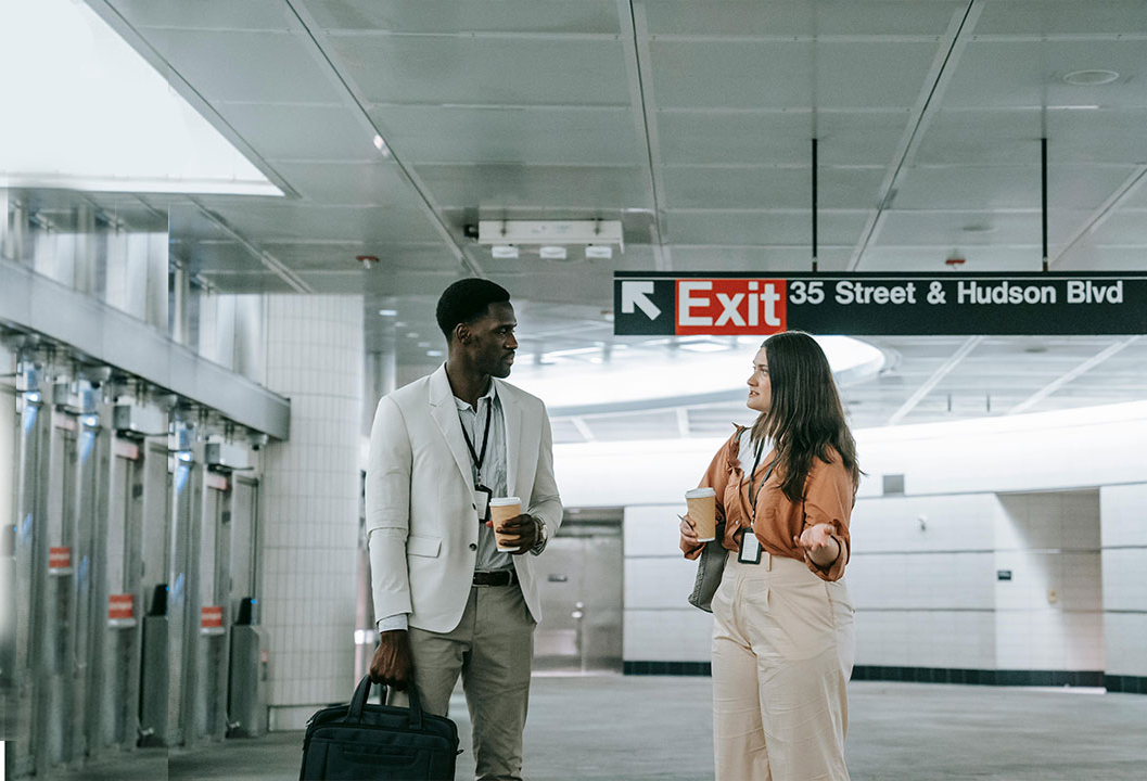 A man and woman holding coffee cups and conversing under a subway exit sign for 35 Street & Hudson Blvd.