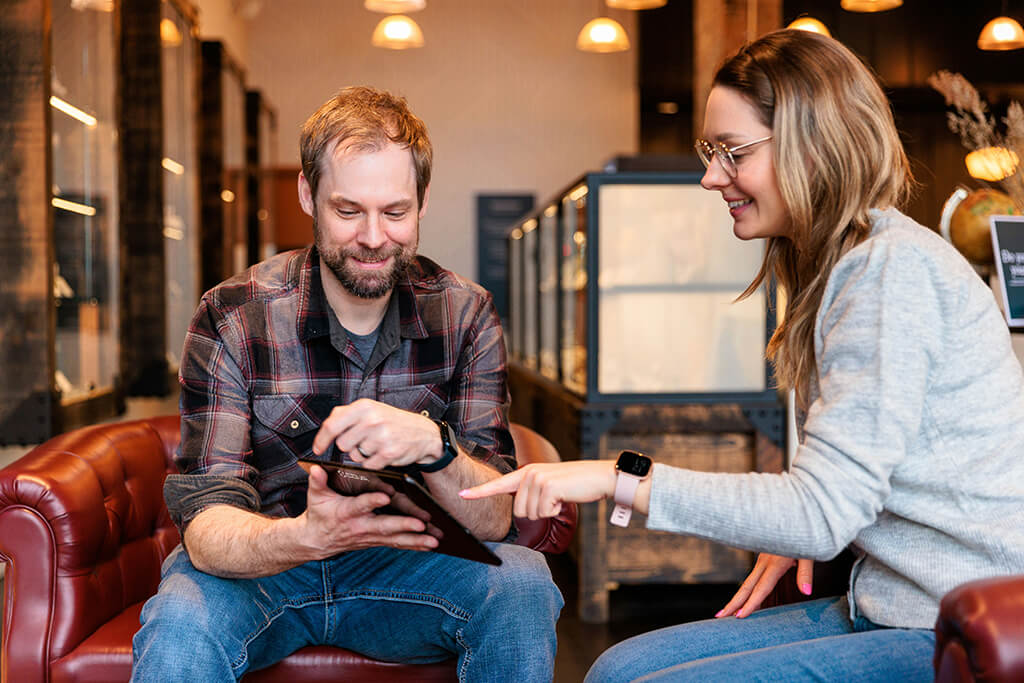 Man and woman sitting on leather chairs indoors, smiling and looking at a tablet the man is holding.