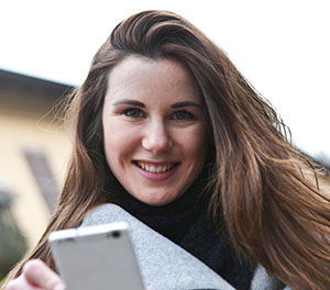 Smiling woman with long brown hair holding a smartphone outdoors.