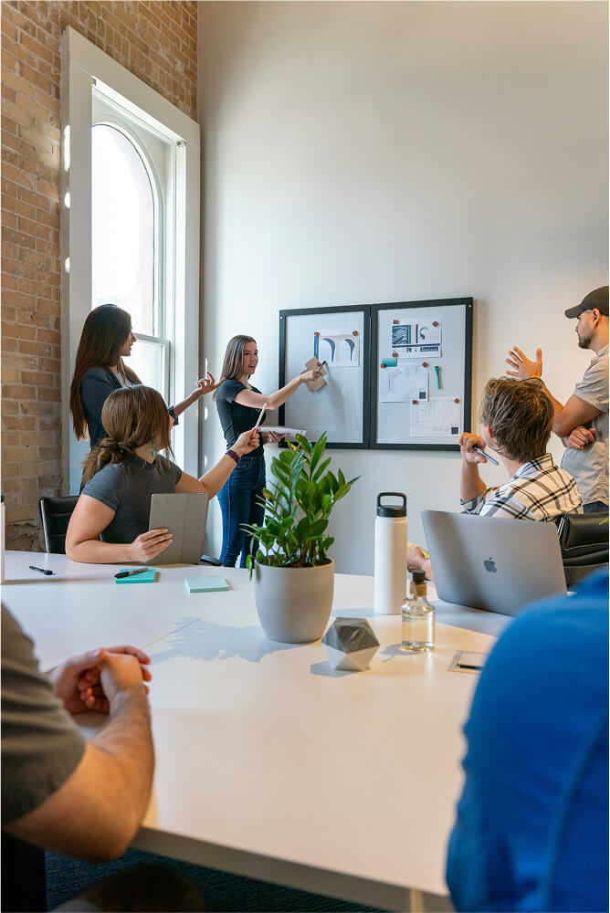 A group of six people in a meeting room discussing and pointing at pinned documents on a wall near a large window.