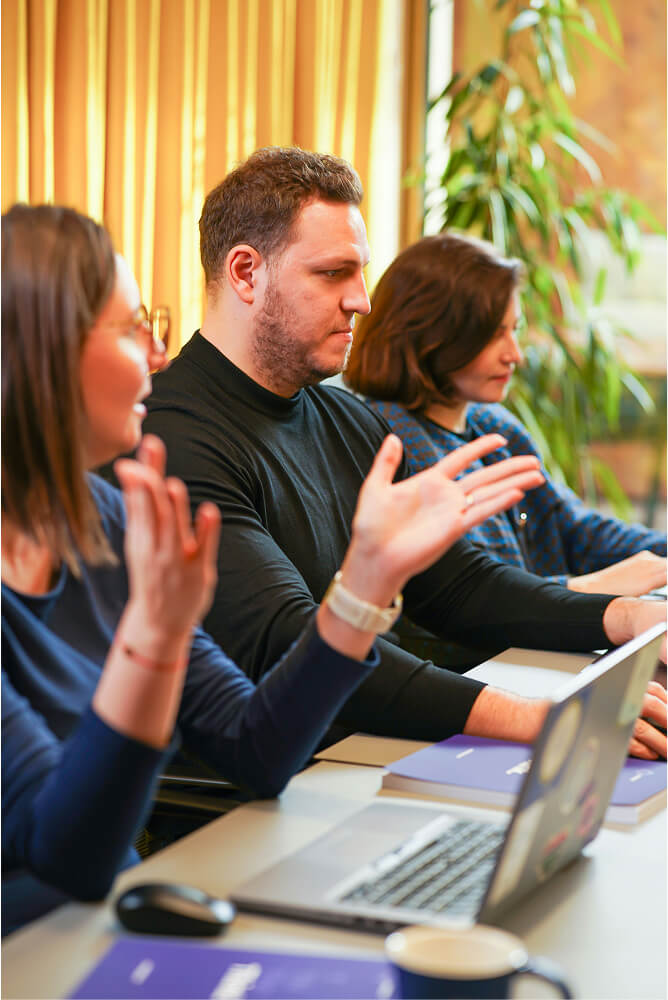 Three people sitting at a table with laptops and notebooks, one person gesturing while speaking.