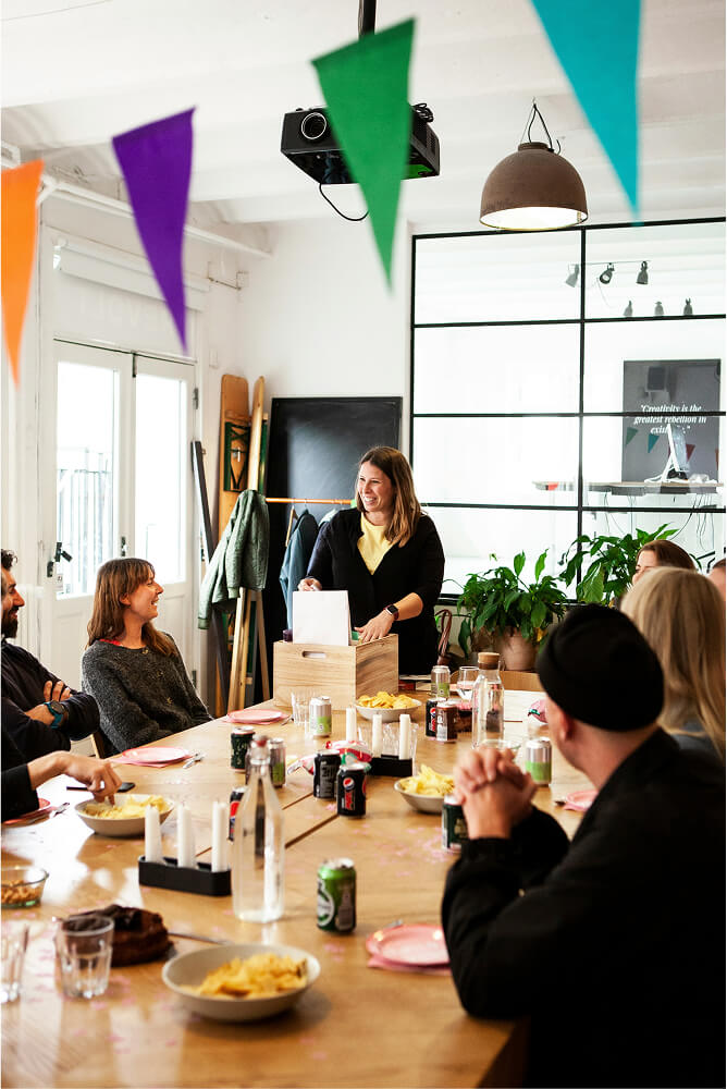 Group of people sitting around a table enjoying snacks and drinks with a woman standing and smiling near a wooden box in a brightly lit room with colorful bunting above.