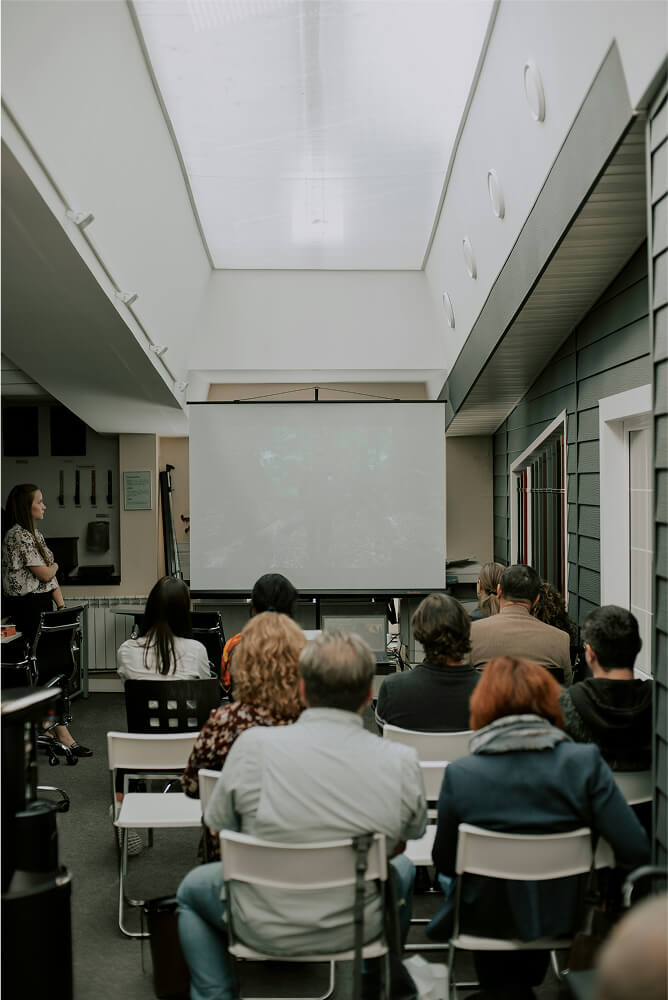 People seated facing a blank projection screen in a modern indoor meeting or classroom setting.