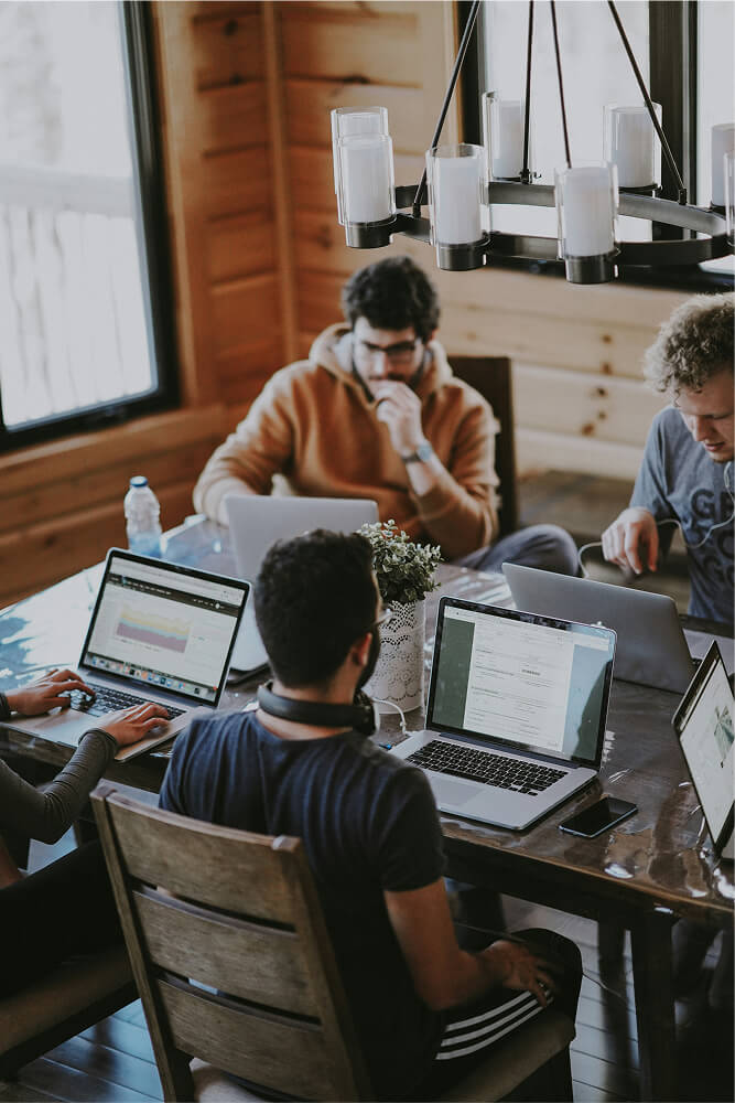 Four people working on laptops around a wooden table in a cozy room with wooden walls and large windows.