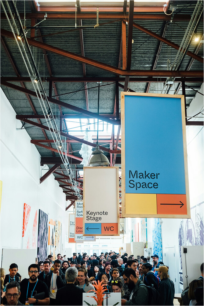 Crowded indoor event with hanging signs indicating directions to Maker Space, Keynote Stage, and WC under high ceiling with exposed beams.