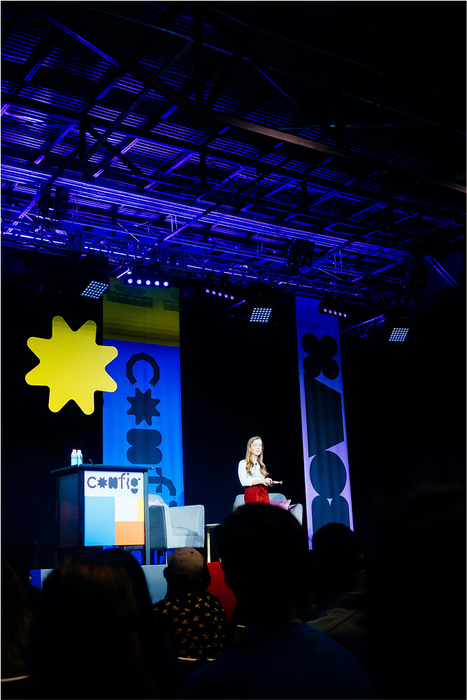 Woman in white top and red skirt speaking on stage at a conference with audience in dark foreground.