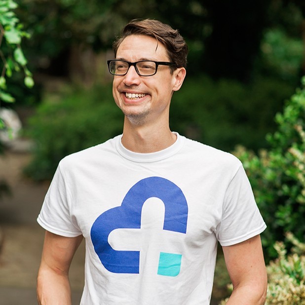 Smiling man with glasses wearing a white t-shirt with a blue and turquoise cloud logo, standing outdoors with green foliage background.