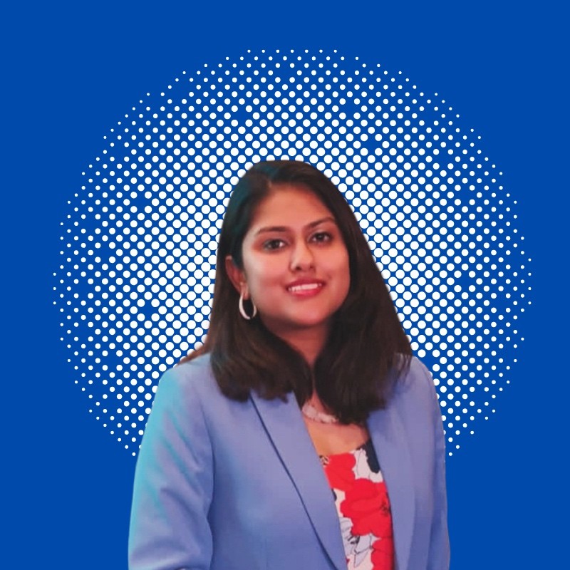 Smiling woman with medium-length dark hair wearing a light blue blazer and floral top, with a white dotted circular pattern on a blue background.