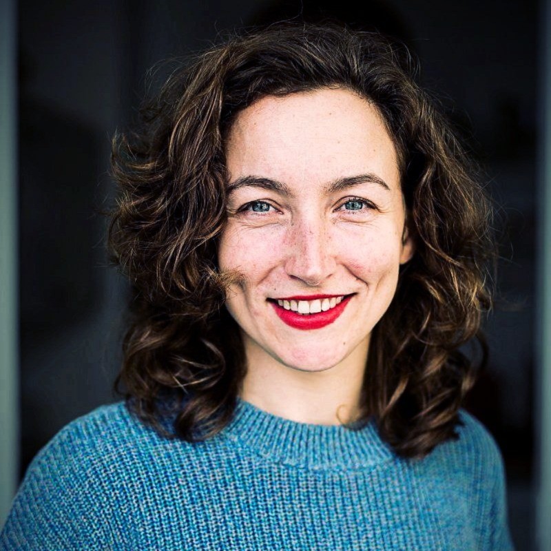 Smiling woman with curly brown hair wearing a blue sweater and bright red lipstick.